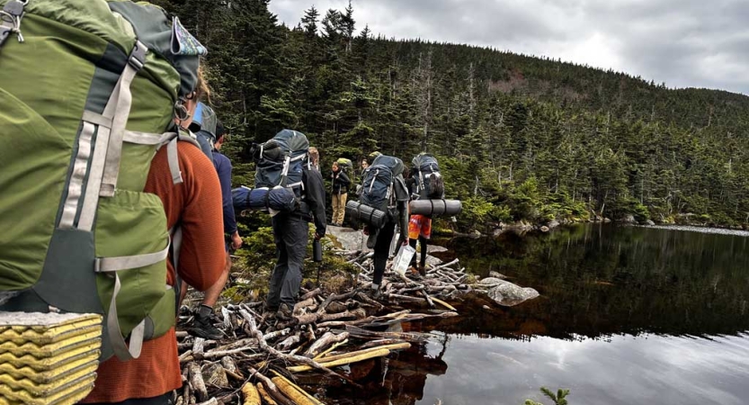 A group of people wearing backpacks hike alongside a still body of water framed by evergreen trees. 